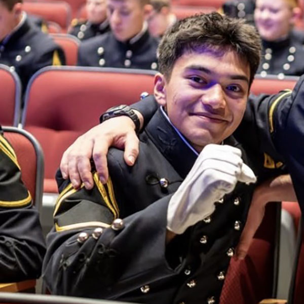A cadet in a formal military uniform sits in an auditorium seat, smiling at the camera while raising a gloved hand in a friendly gesture