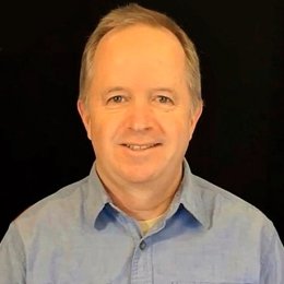 Man wearing a light blue button-up shirt, smiling against a black background.