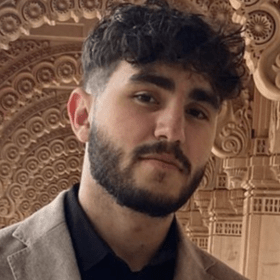 Majd Khalaf with a beard and curly hair, wearing a jacket, posing in front of an ornate architectural background.