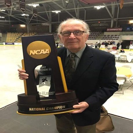 A man in a suit stands with the NCAA hockey trophy on an ice rink.