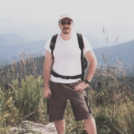 A man stands in hiking gear atop a mountain with a scenic view in the background.