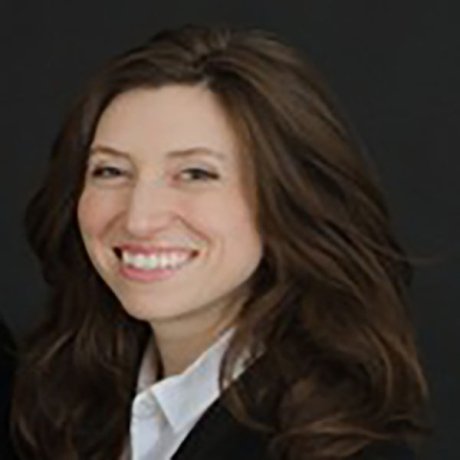 Person with long brown hair wearing a white shirt and dark jacket smiles against a dark background.