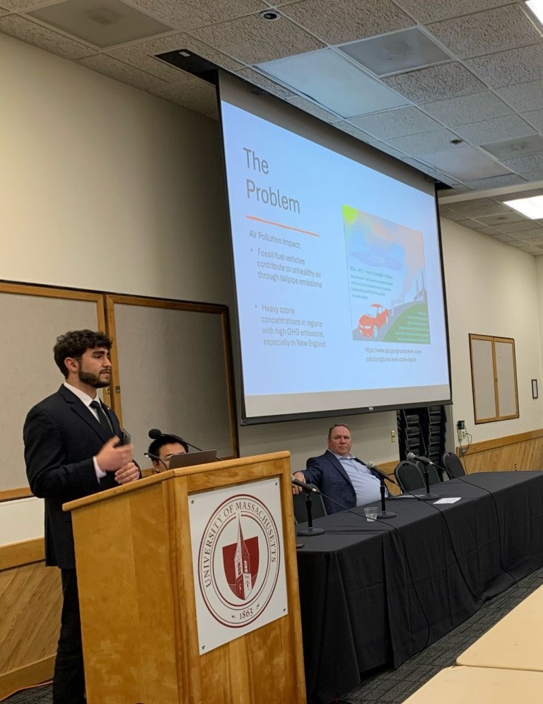 Majd Khalaf presents at a podium with a slide titled "The Problem" projected behind them, discussing air pollution impact and food health. Two individuals seated to the right, all in a conference room at University of Massachusetts.