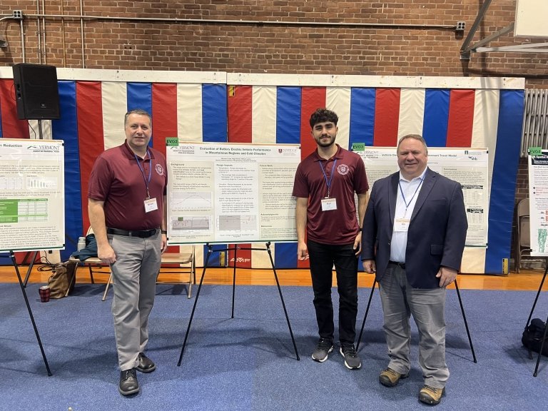 Three individuals standing in front of a display of scientific posters at a conference, flanked by flags hanging in the background.