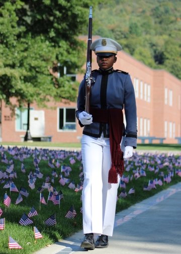 Cadet with a rifle over her shoulder stands along the Tour Strip with flags planted in the grass to her right