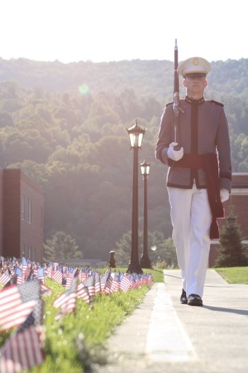 Cadet walks along the Tour Strip with flags planted to his left and a rifle over his shoulder.