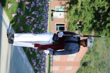 Cadet with a rifle over her shoulder stands along the Tour Strip with flags planted in the grass to her right.